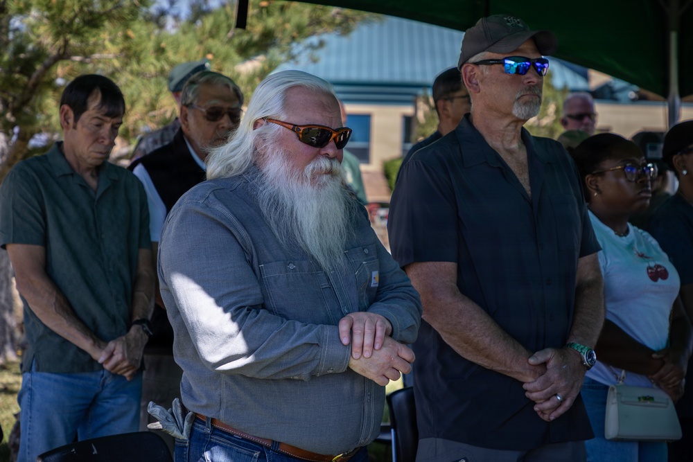 A group of adults standing outdoors at what appears to be a formal or commemorative gathering, possibly a ceremony or memorial event. Several individuals have their hands clasped in front of them in a respectful or attentive posture. The setting includes a shaded canopy area with trees and a building visible in the background.
