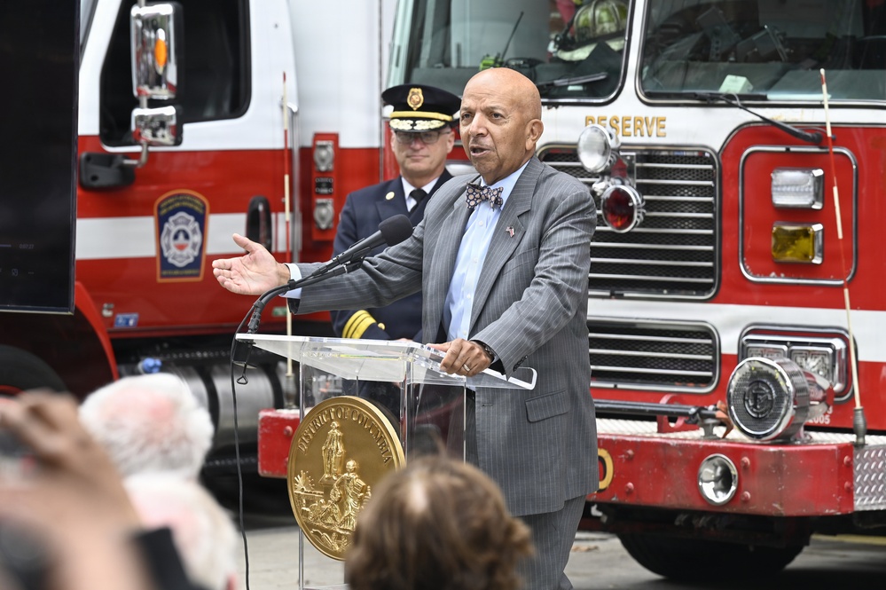 A bald man in a gray pinstripe suit and bow tie speaks at a clear acrylic podium bearing the seal of the District of Columbia. The event appears to be an official public ceremony held outdoors in front of red fire trucks. A uniformed fire official stands in the background behind the speaker.