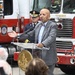 A bald man in a gray pinstripe suit and bow tie speaks at a clear acrylic podium bearing the seal of the District of Columbia. The event appears to be an official public ceremony held outdoors in front of red fire trucks. A uniformed fire official stands in the background behind the speaker.