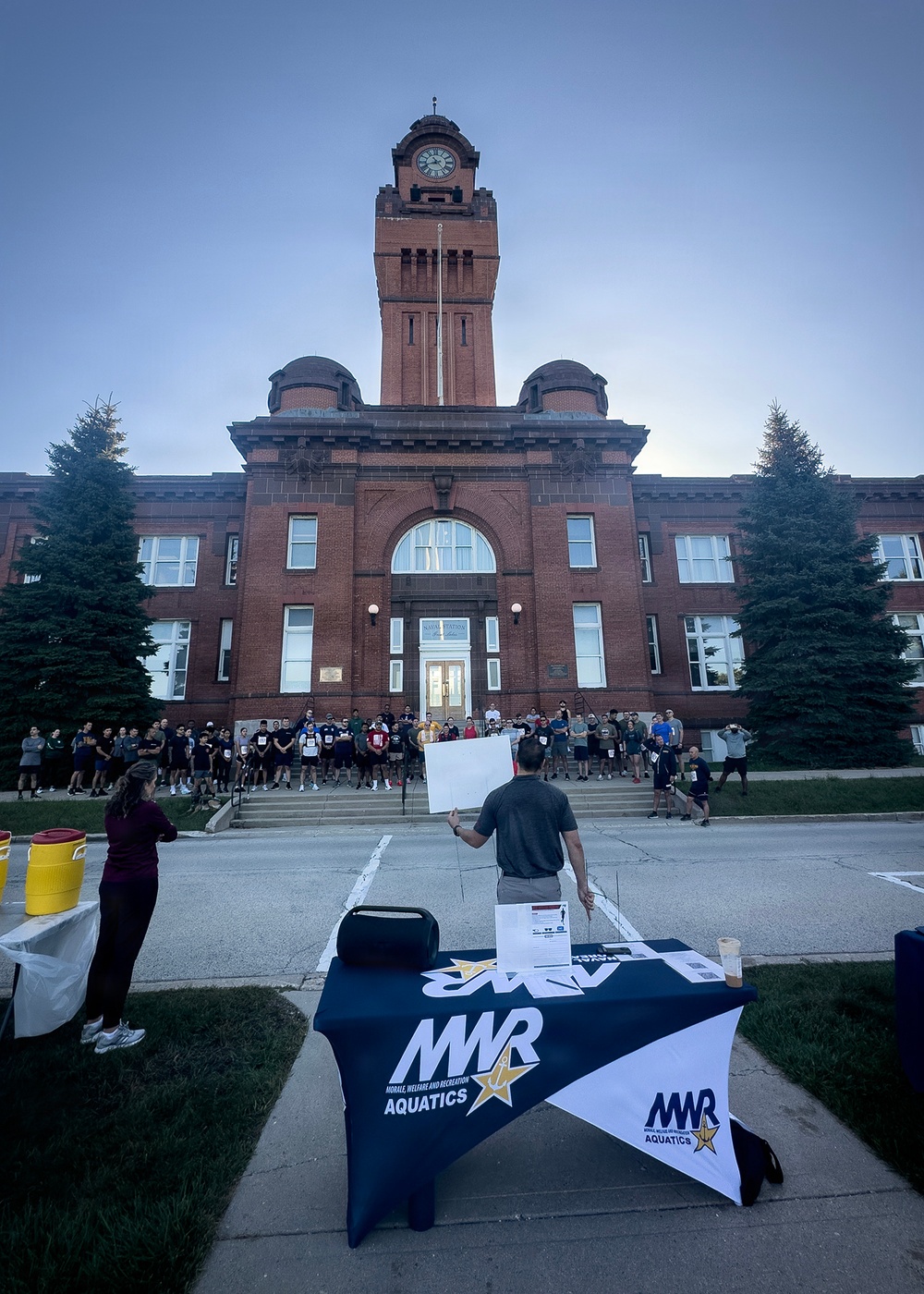 An outdoor event is being held in front of a large red brick building with a clock tower, likely on a military installation. A person stands facing a crowd of participants near an MWR (Morale, Welfare and Recreation) Aquatics branded table, appearing to give instructions or a briefing. The gathering appears to be the start of a run or fitness event.