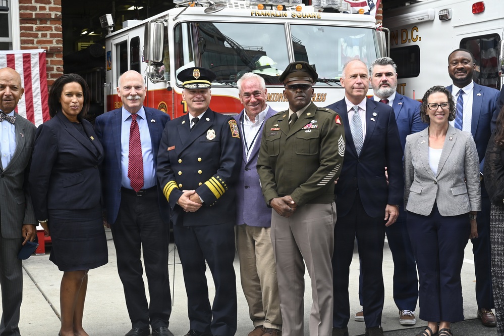 A group of approximately ten officials and dignitaries pose for a formal photograph in front of a fire truck labeled 'Franklin Square' in Washington D.C. The group includes individuals in military dress uniform, fire department command uniform, business attire, and civilian clothing. The setting appears to be a fire station, with an American flag visible in the background.