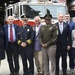A group of approximately ten officials and dignitaries pose for a formal photograph in front of a fire truck labeled 'Franklin Square' in Washington D.C. The group includes individuals in military dress uniform, fire department command uniform, business attire, and civilian clothing. The setting appears to be a fire station, with an American flag visible in the background.