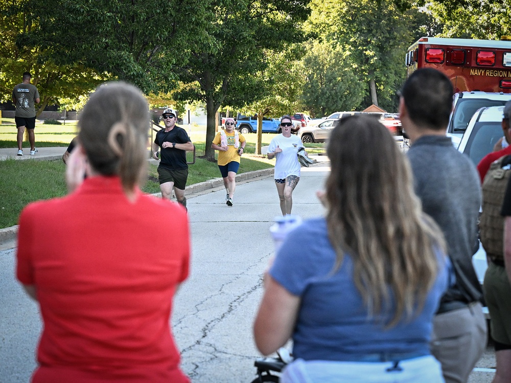 A group of runners approaches spectators along a tree-lined road during what appears to be an organized running event on a military installation. Spectators line the sides of the road cheering on the participants. A red emergency vehicle marked 'Navy Region M...' is visible in the background, suggesting this event is taking place on a U.S. Navy base.
