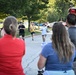 A group of runners approaches spectators along a tree-lined road during what appears to be an organized running event on a military installation. Spectators line the sides of the road cheering on the participants. A red emergency vehicle marked 'Navy Region M...' is visible in the background, suggesting this event is taking place on a U.S. Navy base.