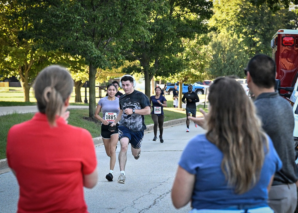 An outdoor running race or fun run is in progress along a tree-lined path. Participants wearing numbered bibs from 'MWR' (Morale, Welfare, and Recreation) are running toward spectators who are cheering them on. The setting appears to be a military installation or park during morning or late afternoon hours based on the lighting.