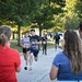 An outdoor running race or fun run is in progress along a tree-lined path. Participants wearing numbered bibs from 'MWR' (Morale, Welfare, and Recreation) are running toward spectators who are cheering them on. The setting appears to be a military installation or park during morning or late afternoon hours based on the lighting.