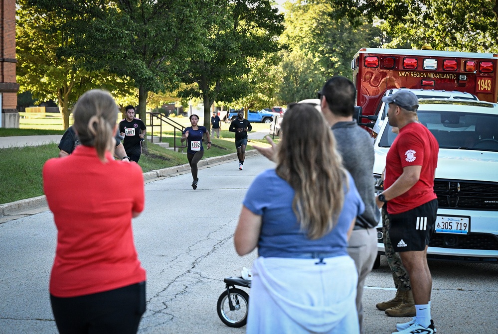 A running event is taking place on a tree-lined road at what appears to be a Navy Region Mid-Atlantic military installation. Three runners wearing race bibs (numbers 1883, 1882, and an unreadable number) are approaching spectators and officials near the finish area. A Navy Region Mid-Atlantic ambulance (unit 1943) is staged nearby, and spectators are cheering the runners on.
