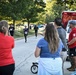 A running event is taking place on a tree-lined road at what appears to be a Navy Region Mid-Atlantic military installation. Three runners wearing race bibs (numbers 1883, 1882, and an unreadable number) are approaching spectators and officials near the finish area. A Navy Region Mid-Atlantic ambulance (unit 1943) is staged nearby, and spectators are cheering the runners on.