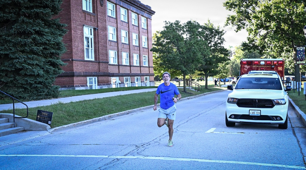 A middle-aged man in a blue Nike shirt and gray shorts is running along a road on what appears to be a military installation, likely a Navy base. A white Dodge Durango SUV with 'Navy Region Mid-Atlantic' markings is parked nearby, and an ambulance is visible in the background. The setting includes a large historic red-brick building and mature trees lining the street.
