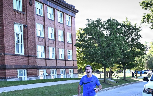 A middle-aged man in a blue Nike shirt and gray shorts is running along a road on what appears to be a military installation, likely a Navy base. A white Dodge Durango SUV with 'Navy Region Mid-Atlantic' markings is parked nearby, and an ambulance is visible in the background. The setting includes a large historic red-brick building and mature trees lining the street.