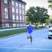A middle-aged man in a blue Nike shirt and gray shorts is running along a road on what appears to be a military installation, likely a Navy base. A white Dodge Durango SUV with 'Navy Region Mid-Atlantic' markings is parked nearby, and an ambulance is visible in the background. The setting includes a large historic red-brick building and mature trees lining the street.