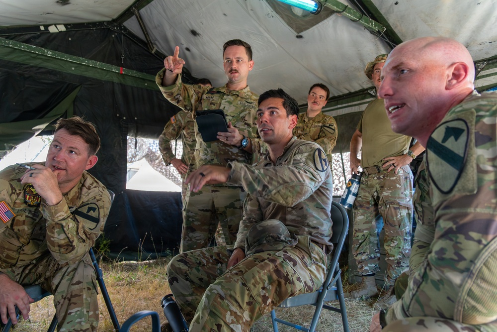 A group of U.S. Army soldiers in MultiCam/OCP camouflage uniforms are engaged in what appears to be a tactical briefing or discussion inside a military field tent. Several soldiers are gesturing and speaking, suggesting an active planning or after-action review session. Visible unit patches indicate affiliation with the 1st Cavalry Division and other Army units.