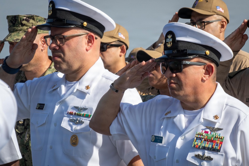 Two U.S. Navy senior enlisted personnel in white service dress uniforms are rendering a salute during a formal military ceremony. Both wear combination covers with 'USN' insignia and display multiple service ribbons and warfare qualification badges. Additional military personnel in various uniforms are visible saluting in the background.