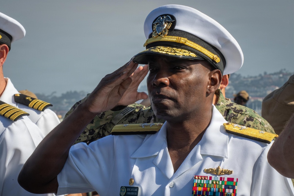 A U.S. Navy flag officer in white service dress uniform is rendering a salute at what appears to be an outdoor military ceremony. The officer wears a name tag and displays multiple rows of service ribbons along with a warfare qualification badge. Several other military personnel in various uniforms are visible in the background.