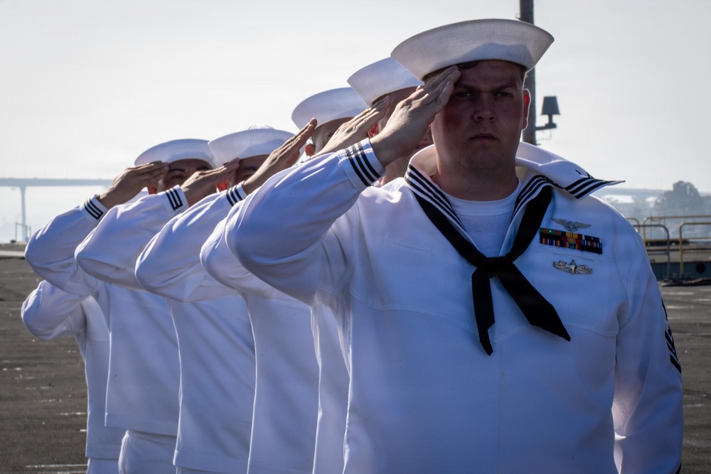 A line of U.S. Navy sailors in dress white uniforms are rendering a formal salute during what appears to be a military ceremony. The foreground sailor displays multiple service ribbons, qualification badges, and a black neckerchief consistent with enlisted dress whites. The setting appears to be on or near a naval vessel or pier, with water visible in the background.