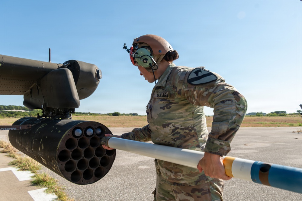 A U.S. Army soldier is loading or inspecting a rocket pod mounted on a military helicopter on an airfield. The soldier is wearing full combat uniform with a flight helmet and hearing protection, and is handling what appears to be a 2.75-inch (70mm) Hydra rocket into a M261 or similar rocket launcher pod. The setting appears to be an outdoor military airfield under clear blue skies.