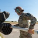 A U.S. Army soldier is loading or inspecting a rocket pod mounted on a military helicopter on an airfield. The soldier is wearing full combat uniform with a flight helmet and hearing protection, and is handling what appears to be a 2.75-inch (70mm) Hydra rocket into a M261 or similar rocket launcher pod. The setting appears to be an outdoor military airfield under clear blue skies.