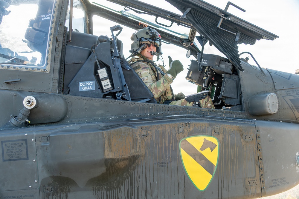 A U.S. Army pilot is seated in the cockpit of a military helicopter, wearing full flight gear including a helmet with visor and giving a thumbs-up gesture. The aircraft bears the distinctive yellow and black shield insignia of the 1st Cavalry Division. The image is taken at close range, clearly showing cockpit details, warning labels, and avionics equipment.