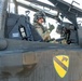 A U.S. Army pilot is seated in the cockpit of a military helicopter, wearing full flight gear including a helmet with visor and giving a thumbs-up gesture. The aircraft bears the distinctive yellow and black shield insignia of the 1st Cavalry Division. The image is taken at close range, clearly showing cockpit details, warning labels, and avionics equipment.