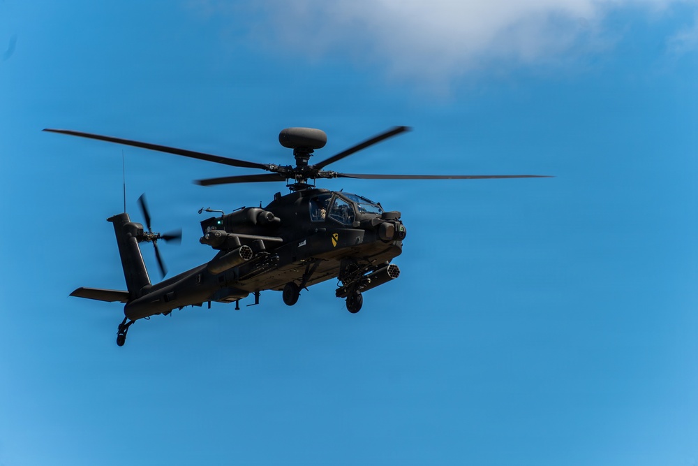 A military attack helicopter is photographed in flight against a clear blue sky with minimal cloud cover. The aircraft is dark/olive colored and appears to be an AH-64 Apache variant, equipped with weapons systems and sensor arrays. The image is taken from a low angle looking upward, providing a detailed view of the helicopter's underside and forward fuselage.