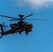 A military attack helicopter is photographed in flight against a clear blue sky with minimal cloud cover. The aircraft is dark/olive colored and appears to be an AH-64 Apache variant, equipped with weapons systems and sensor arrays. The image is taken from a low angle looking upward, providing a detailed view of the helicopter's underside and forward fuselage.