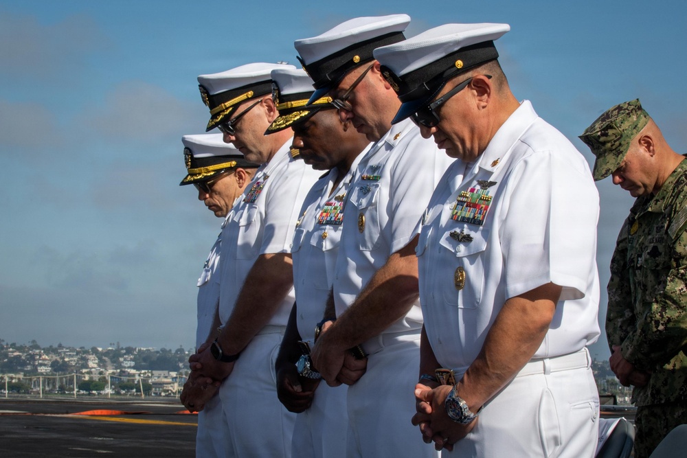 The image shows a group of U.S. Navy officers in white dress uniforms standing with heads bowed in what appears to be a moment of prayer or solemn reflection during a formal military ceremony. They are positioned outdoors, likely on the deck of a naval vessel or pier, with a coastal cityscape visible in the background. A sixth individual in Navy Working Uniform (NWU) camouflage is partially visible on the right edge.