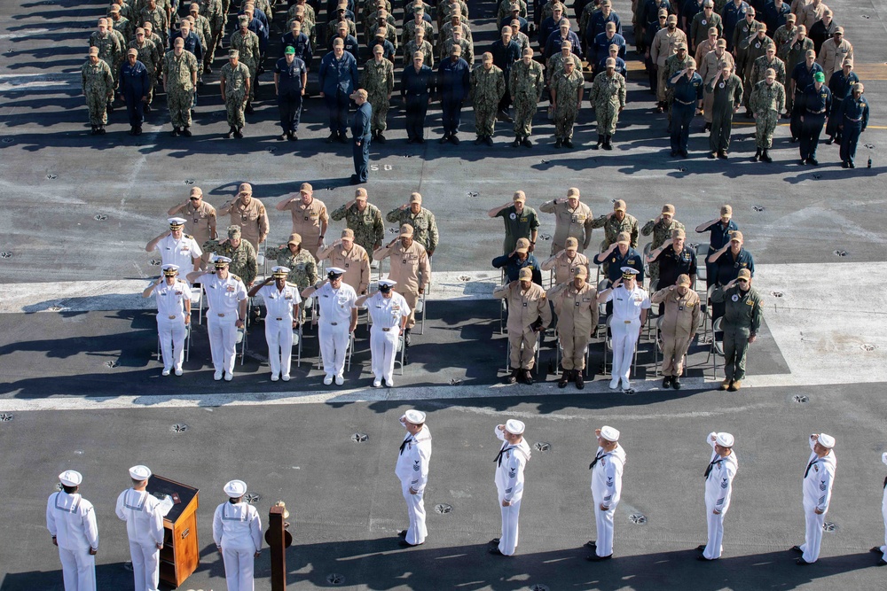 A large military ceremony is taking place on what appears to be the flight deck of an aircraft carrier. Personnel in various uniforms — including white dress uniforms, khaki, camouflage, and flight suits — are assembled in formation, with many individuals rendering salutes. The event appears to be a formal change of command, commissioning, or awards ceremony based on the structured layout and presence of a podium.