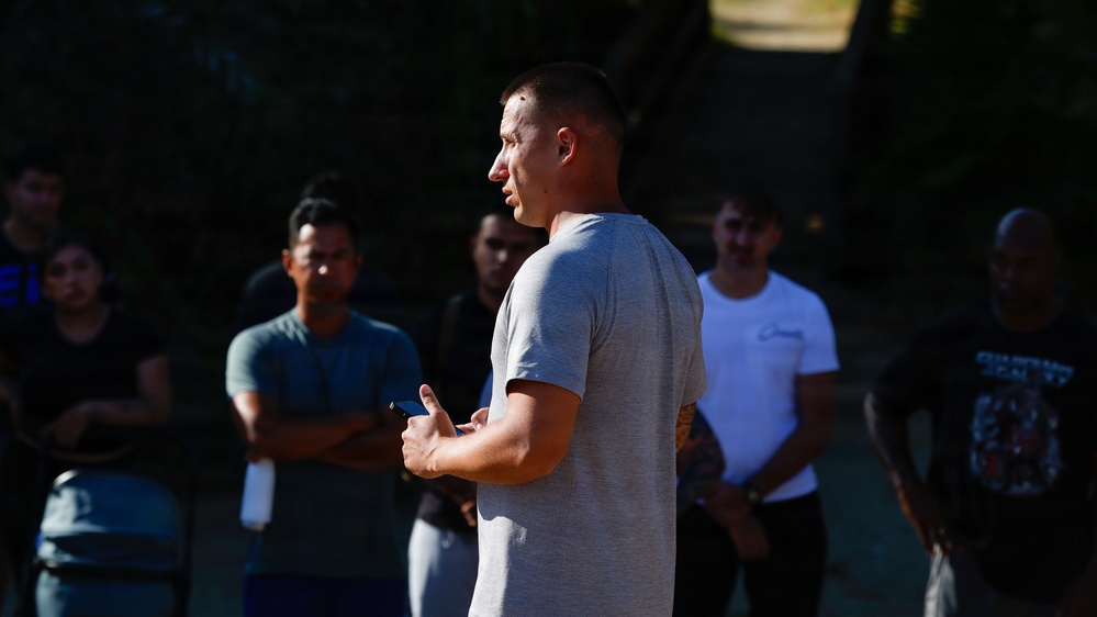 A man in a gray t-shirt stands in the foreground, facing a group of people gathered outdoors, appearing to address or instruct them. He is holding what appears to be a smartphone or small device in his hands. The setting appears to be an outdoor area with trees in the background, with the group listening attentively.