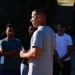 A man in a gray t-shirt stands in the foreground, facing a group of people gathered outdoors, appearing to address or instruct them. He is holding what appears to be a smartphone or small device in his hands. The setting appears to be an outdoor area with trees in the background, with the group listening attentively.