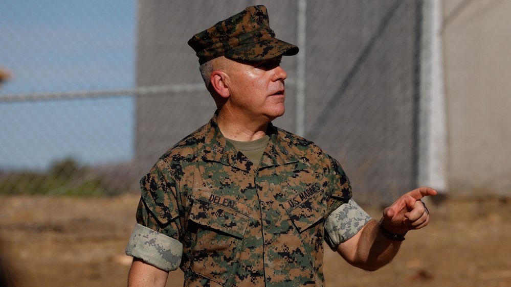 A U.S. Marines servicemember in MARPAT woodland camouflage uniform is photographed outdoors, appearing to speak or brief someone while gesturing with his right hand. His name tape reads 'DELEAL' and the branch tape reads 'U.S. MARINES'. The background shows a chain-link fence and what appears to be a military or industrial facility.