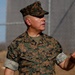 A U.S. Marines servicemember in MARPAT woodland camouflage uniform is photographed outdoors, appearing to speak or brief someone while gesturing with his right hand. His name tape reads 'DELEAL' and the branch tape reads 'U.S. MARINES'. The background shows a chain-link fence and what appears to be a military or industrial facility.