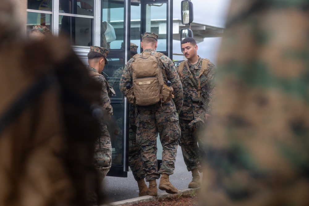 Several military personnel in camouflage uniforms are gathered near the door of a bus or military transport vehicle. The soldiers appear to be in the process of boarding or preparing to depart. At least one soldier is wearing a tan tactical backpack with a visible name tape reading 'SAMUEL'.