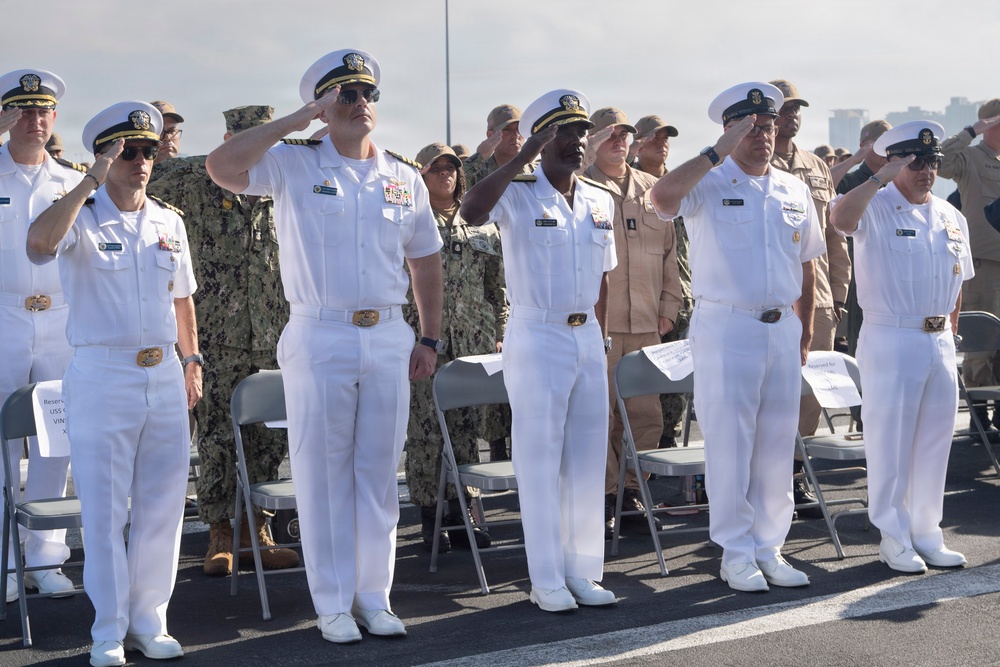 A formal military ceremony featuring U.S. Navy personnel in white dress uniforms rendering salutes. Multiple officers and enlisted members are standing at attention in the foreground, with a larger crowd of personnel in various uniforms visible in the background. The setting appears to be an outdoor venue, likely a pier or naval installation.