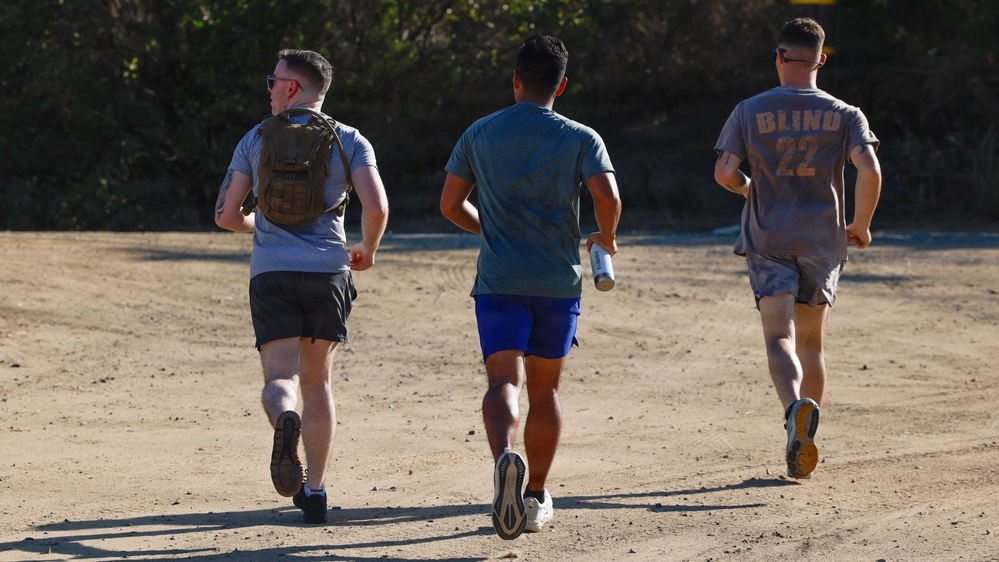 Three men are jogging away from the camera on a dirt path or unpaved surface. The leftmost individual is wearing a tactical/military-style backpack and sunglasses, the center person is carrying a water bottle, and the rightmost person is wearing a gray shirt with 'BLIND 22' printed on the back. The setting appears to be an outdoor trail or training area with vegetation visible in the background.