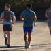 Three men are jogging away from the camera on a dirt path or unpaved surface. The leftmost individual is wearing a tactical/military-style backpack and sunglasses, the center person is carrying a water bottle, and the rightmost person is wearing a gray shirt with 'BLIND 22' printed on the back. The setting appears to be an outdoor trail or training area with vegetation visible in the background.