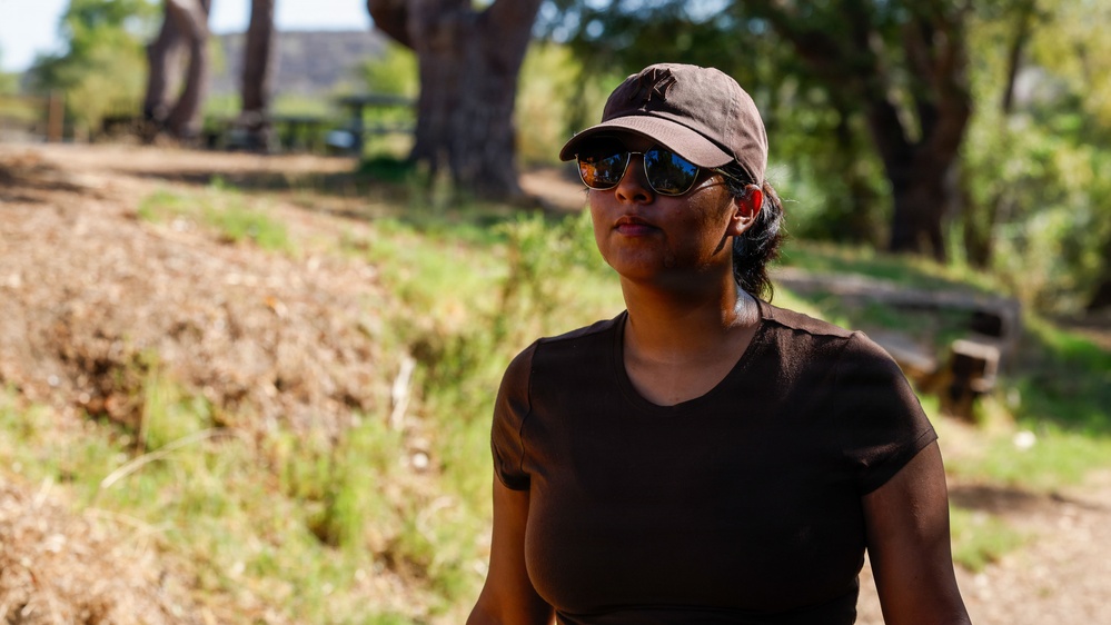 A person stands outdoors in a natural park or trail setting during daylight hours. They are wearing a brown baseball cap, mirrored aviator sunglasses, and a dark brown t-shirt. The background features trees, dry grass, and what appears to be a picnic area.