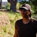 A person stands outdoors in a natural park or trail setting during daylight hours. They are wearing a brown baseball cap, mirrored aviator sunglasses, and a dark brown t-shirt. The background features trees, dry grass, and what appears to be a picnic area.