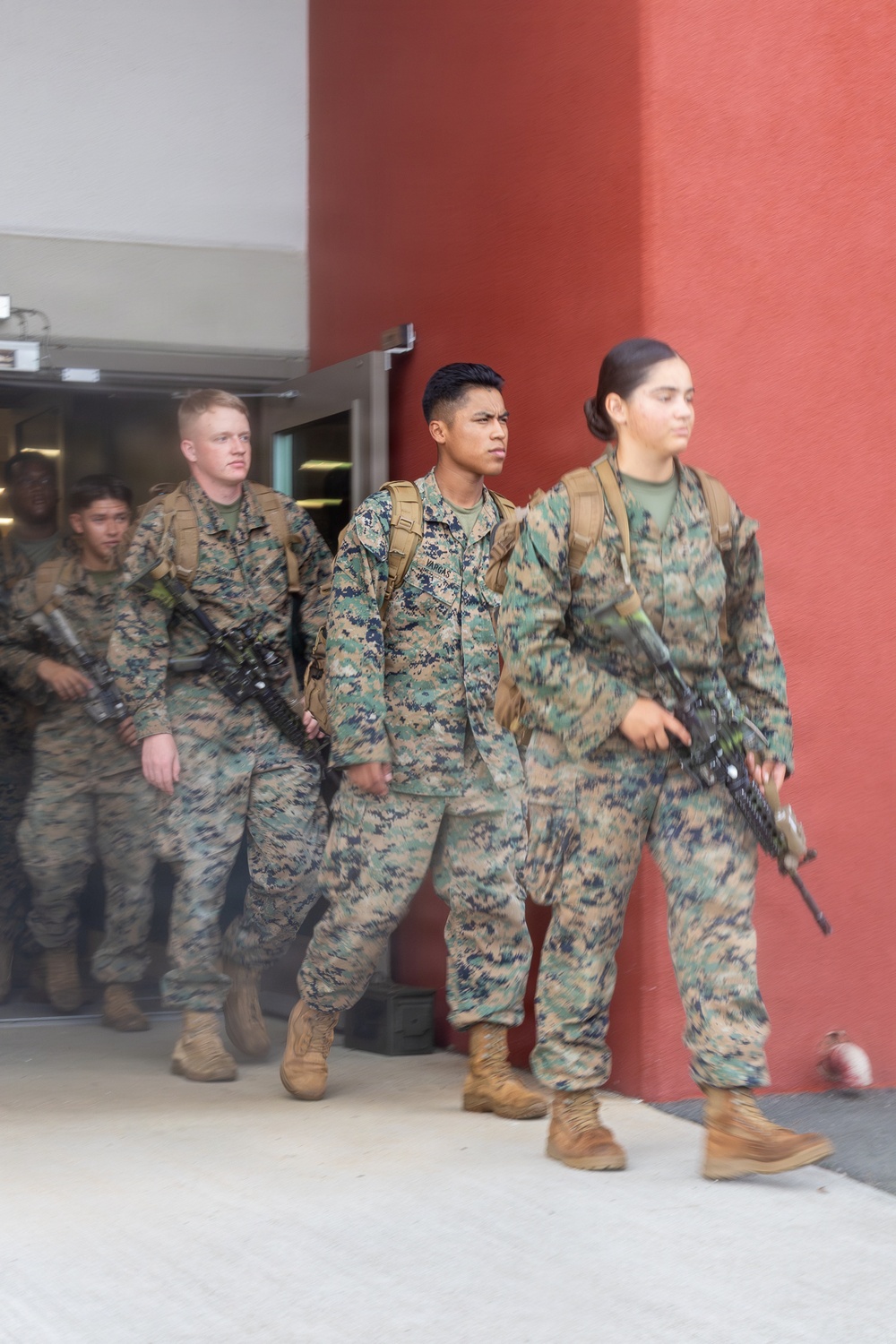 A group of military personnel in MARPAT (Marine pattern) digital camouflage uniforms are exiting a building through a glass door. They are carrying rifles and wearing tactical backpacks, suggesting a training exercise or operational movement. At least one name tape reading 'VARGAS' is partially visible on one of the service members.