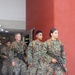 A group of military personnel in MARPAT (Marine pattern) digital camouflage uniforms are exiting a building through a glass door. They are carrying rifles and wearing tactical backpacks, suggesting a training exercise or operational movement. At least one name tape reading 'VARGAS' is partially visible on one of the service members.