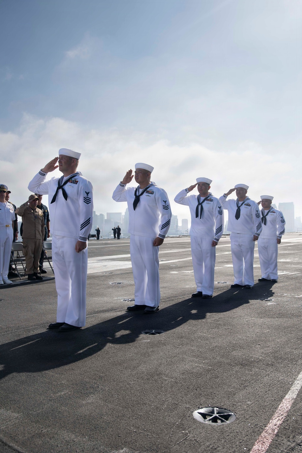 A formal U.S. Navy ceremony is taking place on what appears to be the flight deck of an aircraft carrier. Multiple sailors in dress white uniforms are rendering salutes in formation, with a hazy city skyline visible in the background. Officers in khaki uniforms are visible observing from the left side of the frame.
