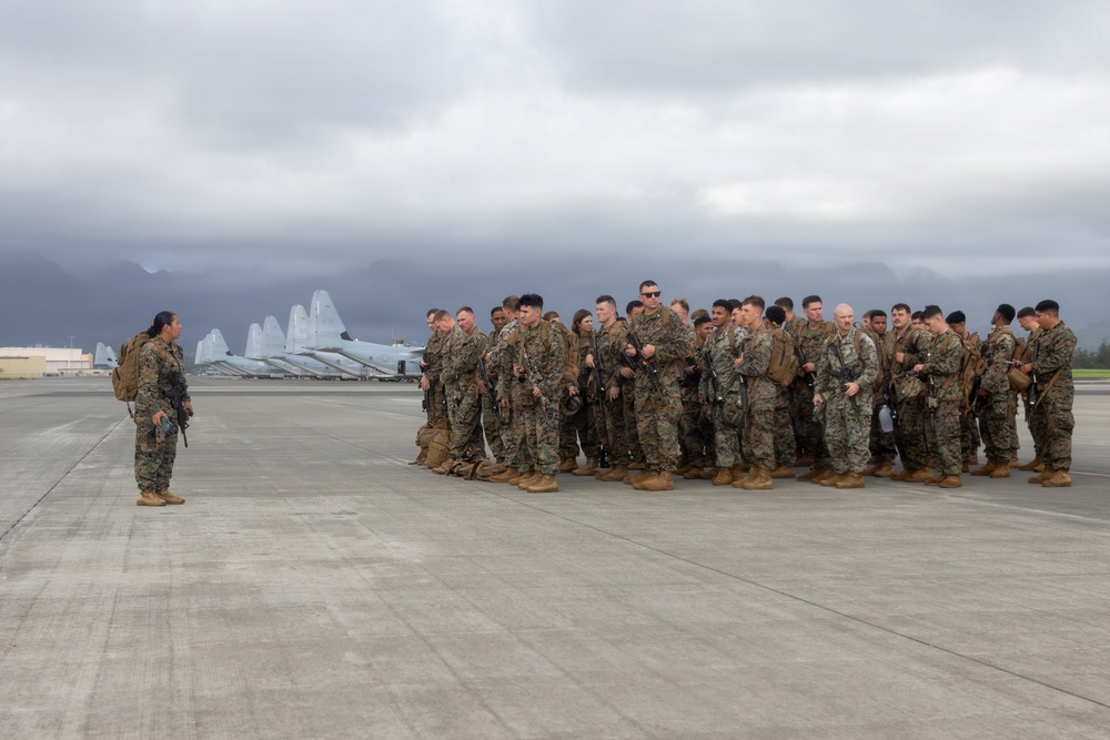 A group of military personnel in camouflage uniforms are assembled on an airfield tarmac, facing a single individual who appears to be addressing or briefing them. Multiple large military aircraft are visible in the background, along with mountainous terrain partially obscured by overcast clouds. The setting suggests a pre-mission briefing or post-arrival assembly at a military air installation.