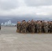 A group of military personnel in camouflage uniforms are assembled on an airfield tarmac, facing a single individual who appears to be addressing or briefing them. Multiple large military aircraft are visible in the background, along with mountainous terrain partially obscured by overcast clouds. The setting suggests a pre-mission briefing or post-arrival assembly at a military air installation.