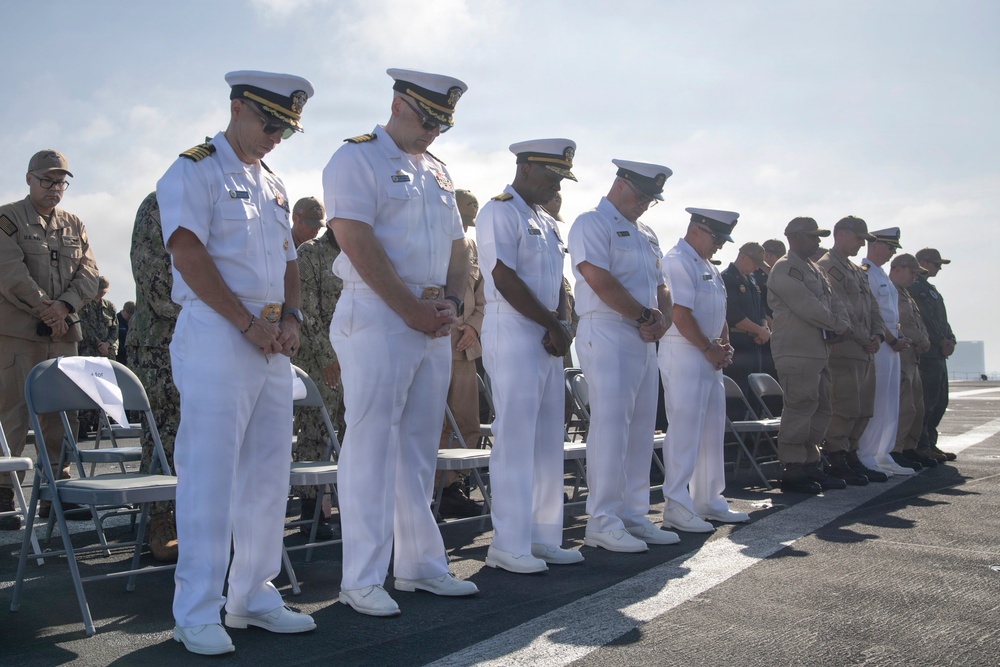 A formal military ceremony is taking place outdoors on what appears to be the flight deck of a naval vessel. Multiple naval officers in white dress uniforms are standing with heads bowed in a moment of prayer or reflection, alongside personnel in other military uniforms. The gathering suggests an official commemorative or change-of-command ceremony.