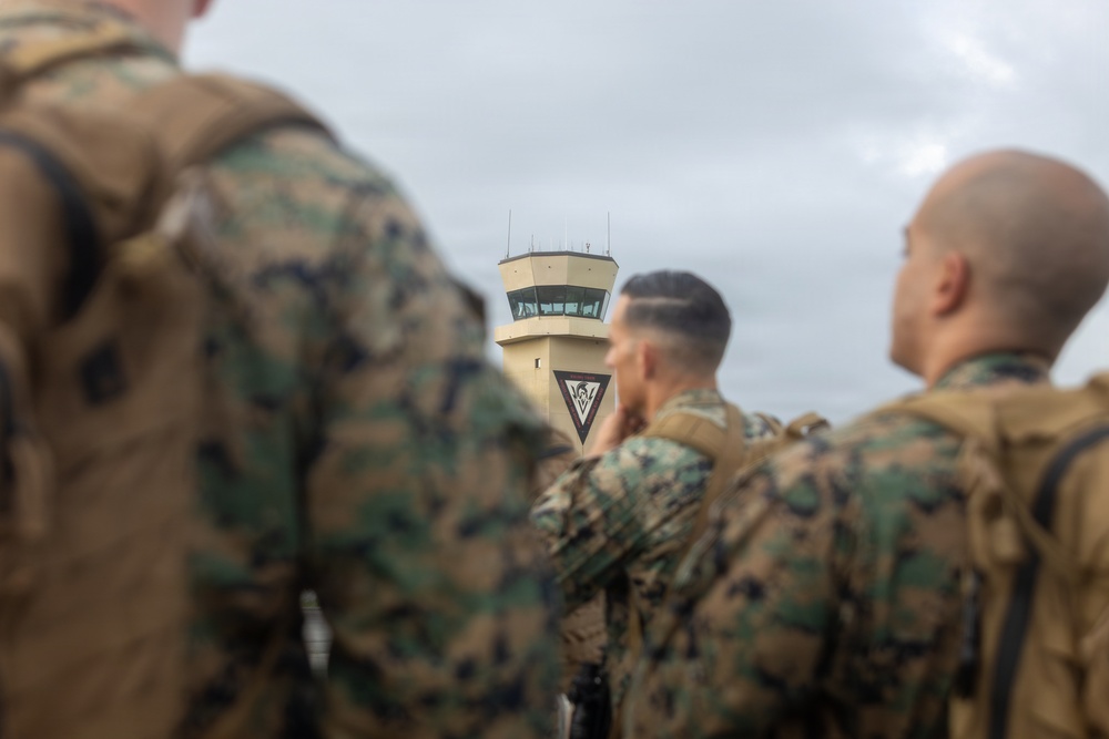 Several military personnel in woodland camouflage uniforms (MARPAT pattern) are gathered on what appears to be an airfield tarmac. In the background, an air traffic control tower is visible with a triangular insignia/emblem displayed on its facade. The foreground figures are out of focus while the control tower remains the sharpest element in the mid-ground.