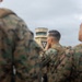 Several military personnel in woodland camouflage uniforms (MARPAT pattern) are gathered on what appears to be an airfield tarmac. In the background, an air traffic control tower is visible with a triangular insignia/emblem displayed on its facade. The foreground figures are out of focus while the control tower remains the sharpest element in the mid-ground.
