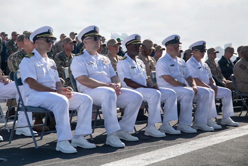 A formal military ceremony featuring several U.S. Navy officers seated in the front row wearing dress white uniforms, with a large crowd of mixed military personnel in various uniforms seated behind them. The setting appears to be an outdoor venue, likely on a naval base or ship deck, suggesting an official commissioning, change of command, or similar ceremonial event. The officers in the front row display various rank insignia and service ribbons on their uniforms.