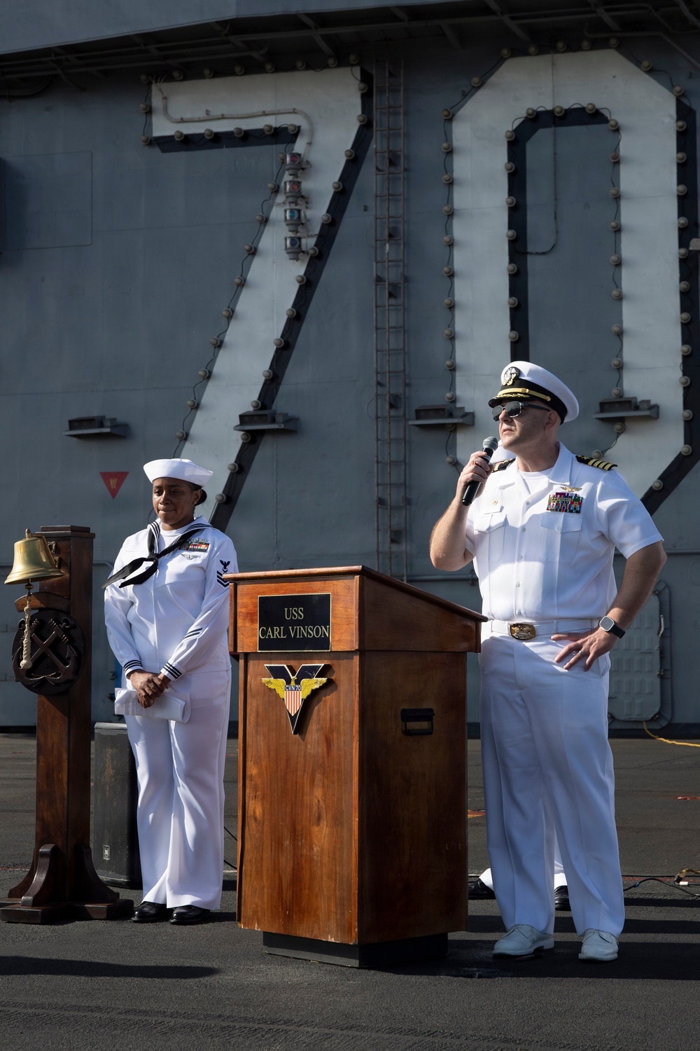 A U.S. Navy officer in dress white uniform speaks at a podium labeled 'USS Carl Vinson' on the flight deck of the aircraft carrier CVN-70. A junior enlisted sailor stands at attention nearby, and the ship's island structure with the hull number '70' is visible in the background. This appears to be a formal ceremony or official event aboard the vessel.
