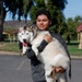 A young man is holding a Siberian Husky dog in his arms at what appears to be a public park. The dog has a gray and white coat with its tongue out, and the man is carrying it with both arms. The setting includes park pavilions, green grass, trees, and a paved path in the background.