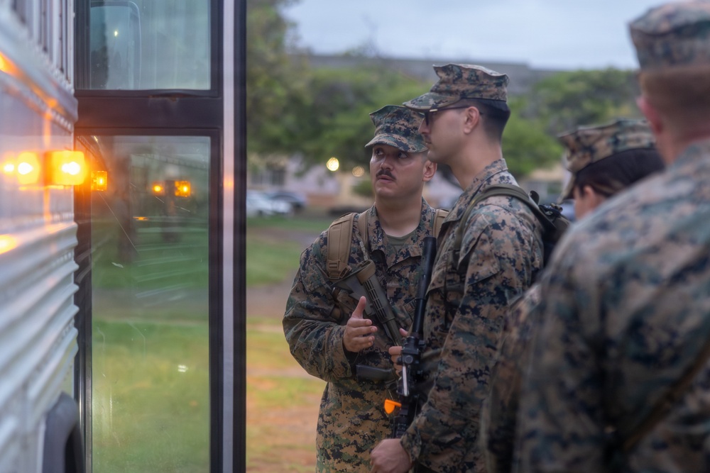 Several U.S. Marines in MARPAT woodland camouflage uniforms and covers are gathered beside a bus with amber warning lights illuminated. The Marines are carrying rifles and wearing tactical backpacks, appearing to be in conversation near what looks like a boarding or staging area. The setting appears to be a military installation during early morning or dusk hours.
