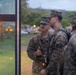Several U.S. Marines in MARPAT woodland camouflage uniforms and covers are gathered beside a bus with amber warning lights illuminated. The Marines are carrying rifles and wearing tactical backpacks, appearing to be in conversation near what looks like a boarding or staging area. The setting appears to be a military installation during early morning or dusk hours.